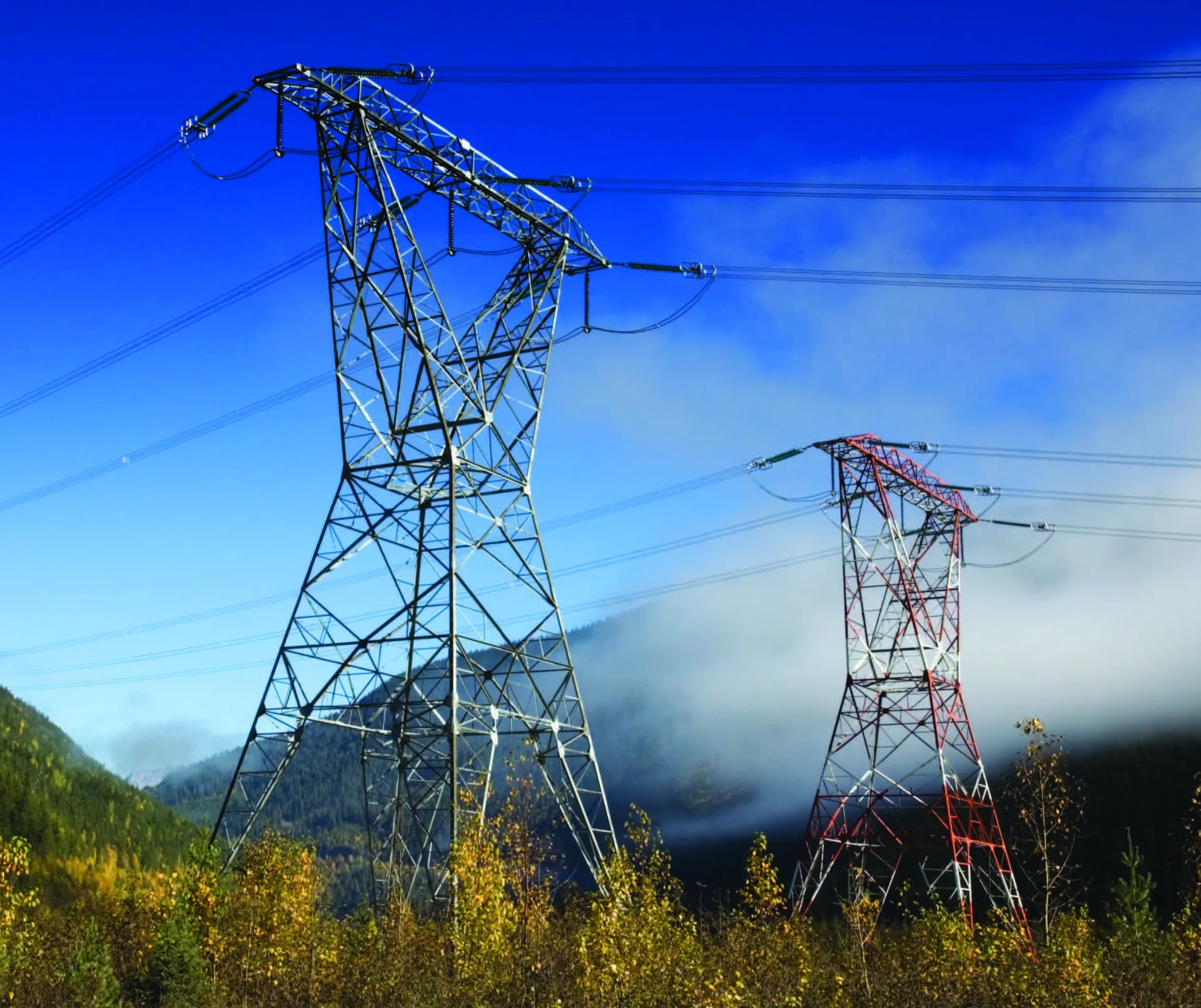 Two lattice transmission towers on a forested mountain slope above low cloud